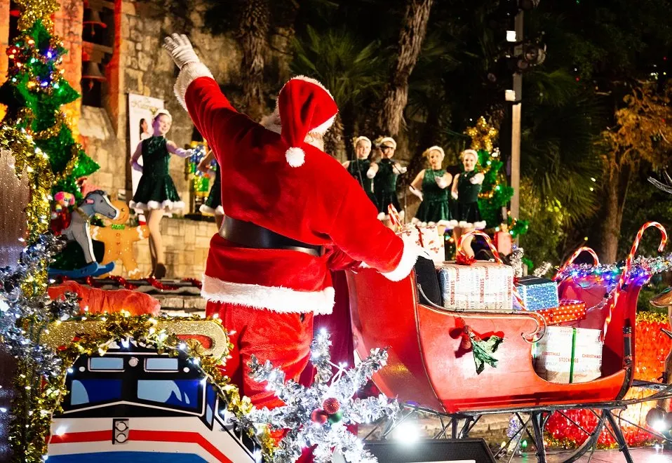 A man dressed as Santa Claus holds his left hand up and pushes a decorated sleigh.
