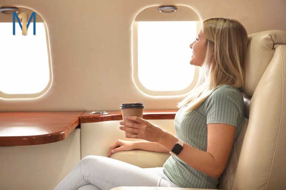 A woman sits in a private jet and looks out the window. She is holding a coffee cup.
